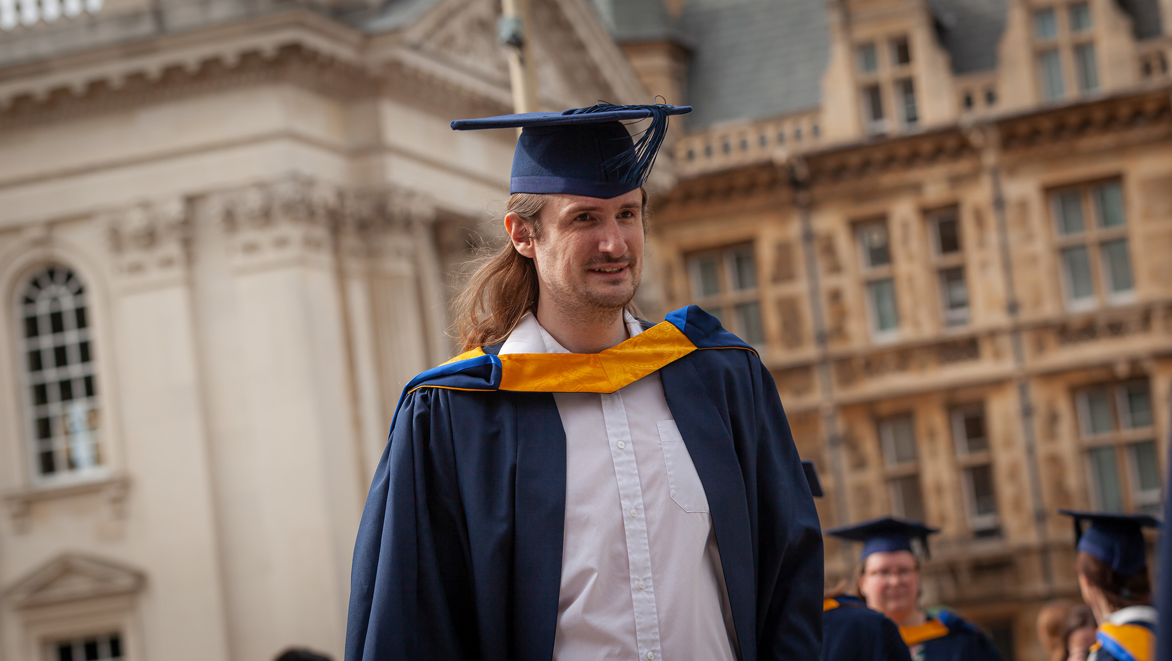 Data scientist graduate, Adam Sanigar, outside the Old Schools building walks toward the Cambridge Corn Exchange dressed in a navy and yellow cap and gown for the graduation ceremony.