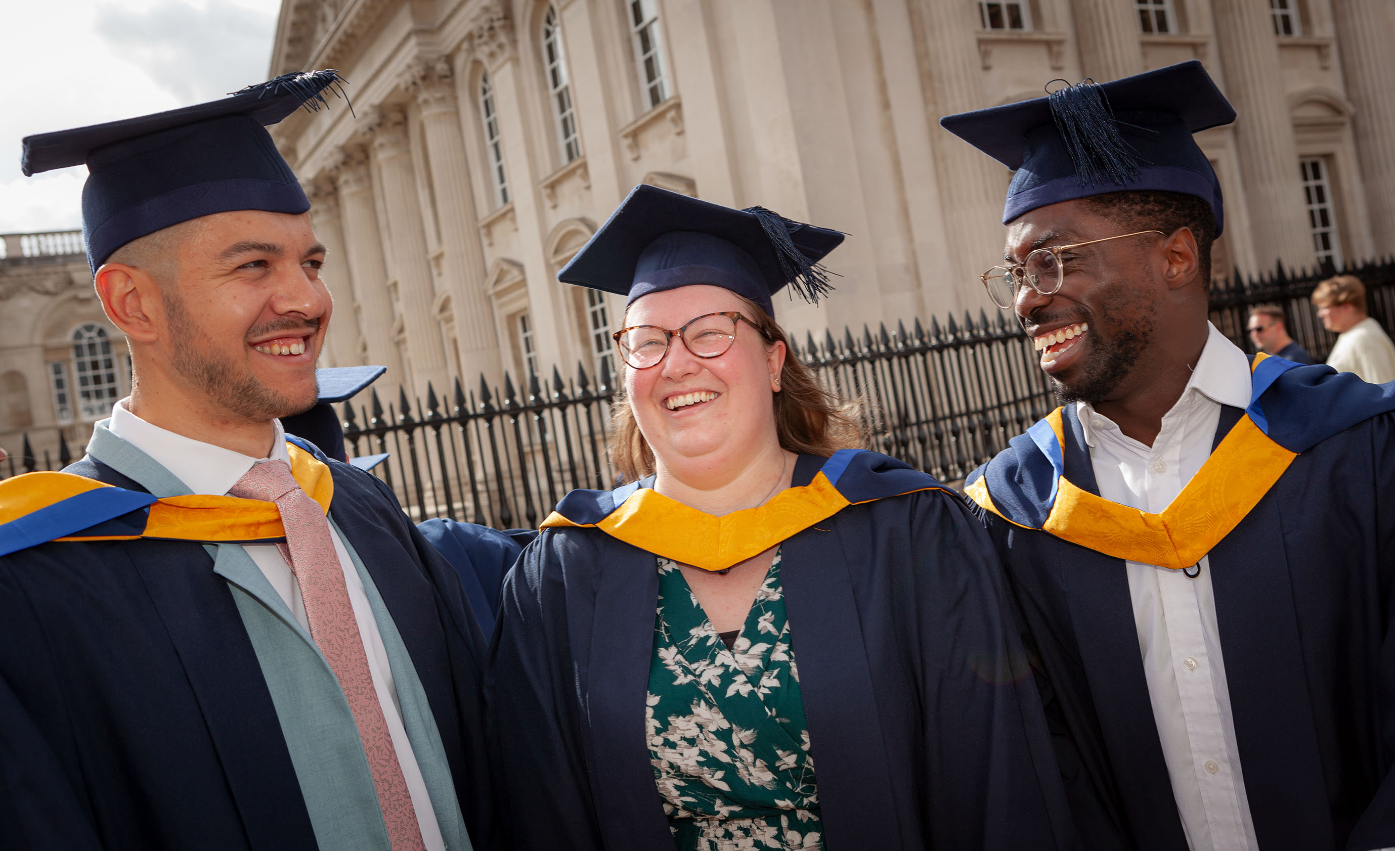 Data scientist apprentice graduates smile outside the graduation ceremony held at the Cambridge Corn Exchange dressed in navy and yellow cap and gowns.