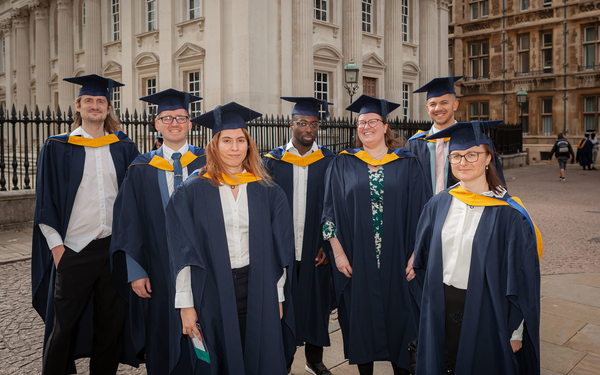 Cambridge's first-ever cohort of data scientist BSc graduates stand and pose outside the Old Schools building dressed in navy and yellow cap and gowns.