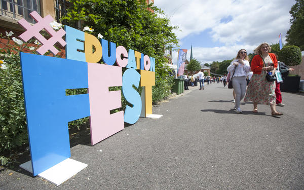 large letter standing on the ground spelling out the phrase hashtag education fest with two smiling festival goers are walking past