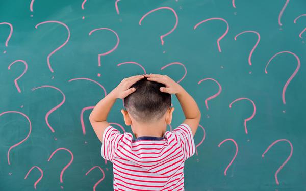 a boy with his hands on his head facing a chalk board covered in question marks