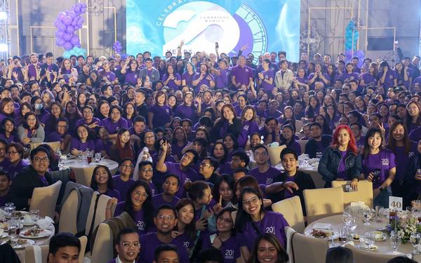 Cambridge's 900 manila colleagues gather together on and off stage in front of a screen for a group photo dressed in Cambridge's purple 20th anniversary t-shirts.